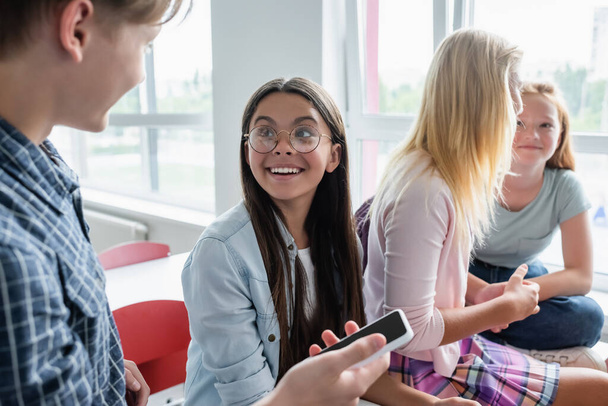 Smiling schoolgirl looking at blurred classmate with smartphone in classroom  - Foto, Imagen