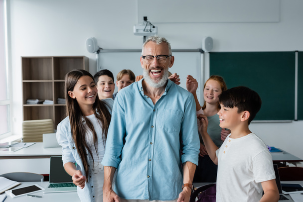 cheerful multiethnic classmates near happy teacher laughing with closed eyes in classroom - Photo, Image