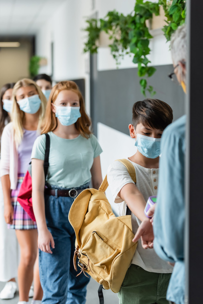 размытый учитель проверяет температуру многонациональных учеников в медицинских масках с помощью пирометра - Фото, изображение