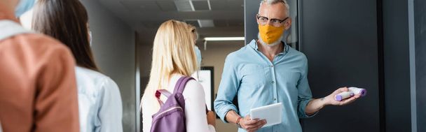 maestro en máscara médica sosteniendo pirómetro mientras señala en el aula cerca de colegiala, pancarta - Foto, imagen