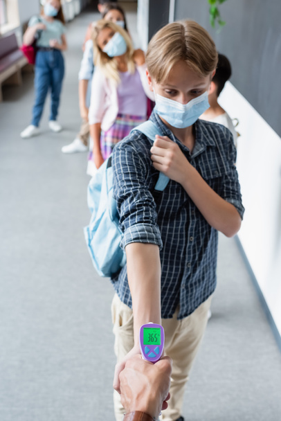 έφηβος με ιατρική μάσκα κοντά στο δάσκαλο μετρώντας τη θερμοκρασία του με πυρομετρητή - Φωτογραφία, εικόνα
