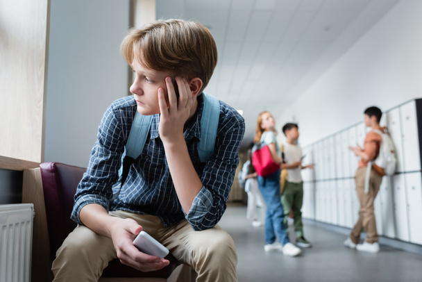 niño deprimido con teléfono inteligente sentado solo en el pasillo de la escuela cerca de los adolescentes en un fondo borroso - Foto, Imagen