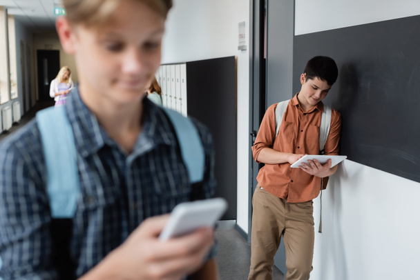 adolescente utilizando tableta digital cerca de compañero de clase con teléfono móvil en primer plano borrosa - Foto, Imagen