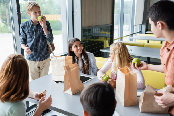 щасливі підлітки їдять бутерброди та яблука під час розмови в шкільній їдальні
 - Фото, зображення