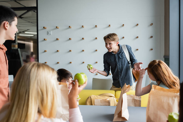 cheerful boy gesturing while talking to classmates in school eatery - Photo, Image