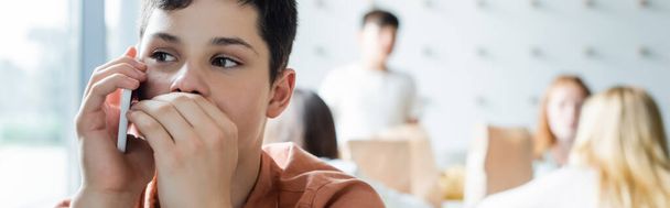 школьник, прикрывающий рот рукой во время разговора по мобильному телефону рядом с размытыми одноклассниками, баннер - Фото, изображение