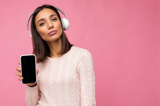 Beautiful happy young woman wearing stylish casual outfit isolated on background wall holding and showing mobile phone with empty display for mockup wearing white bluetooth headphones listening to - Foto, afbeelding