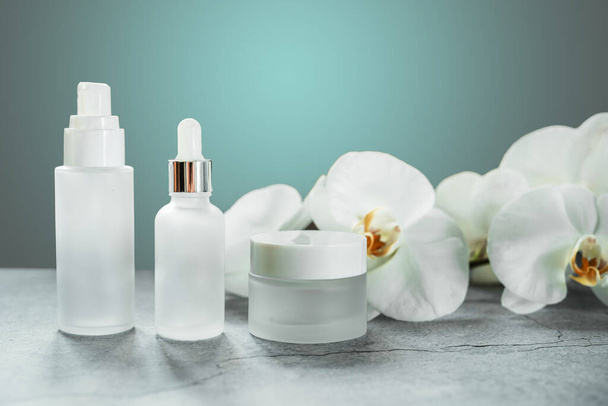 White glass serum bottle and cream jar in the bathroom with orchid flowers in the background, unbranded cosmetic products, spa cosmetic product branding mockup - Фото, изображение