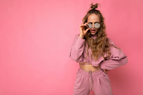 Aantrekkelijke zelfverzekerde positieve jonge blonde vrouw draagt alledaagse stijlvolle kleding en moderne zonnebril geïsoleerd op kleurrijke achtergrond muur kijken naar camera - Foto, afbeelding
