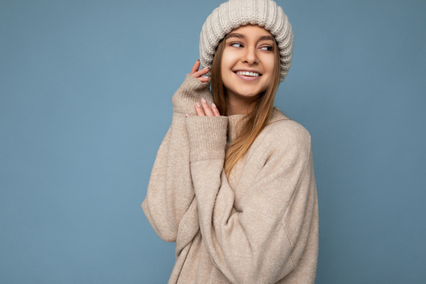Cozy cute photo of beautiful happy smiling young dark blonde female person isolated over blue background wall wearing warm sweater and knitted hat looking to the side - Zdjęcie, obraz