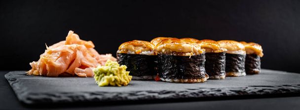 Panoramafoto von serviertem warmem Sushi in Nori-Blättern mit Ingwer und Wasabi auf schwarzem Steinteller. - Foto, Bild