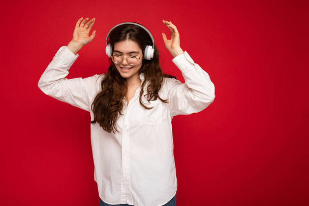 Vonzó boldog mosolygós fiatal barna nő személy visel fehér ing és optikai szemüveg elszigetelt piros háttér visel fehér vezeték nélküli bluetooth fülhallgató zenét hallgatni és táncolni - Fotó, kép