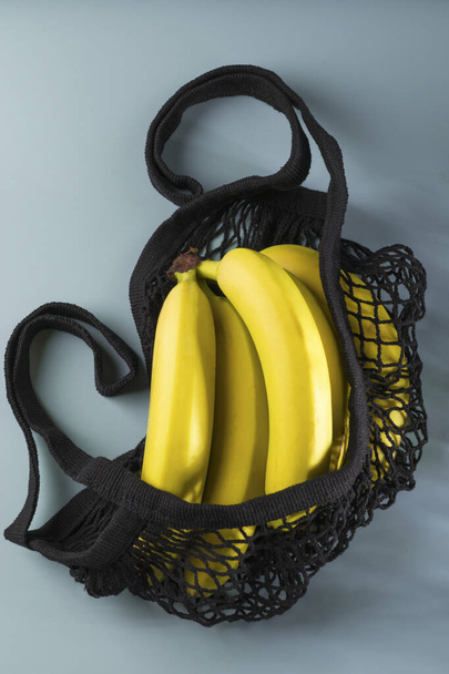 廃棄物ゼロのコンセプト。青い背景に黒のエコ綿のストリングバッグに黄色の熟したバナナ。再利用可能なメッシュショッピングバッグ。平置きだ。鍵が低い。上からの眺め。垂直位置 - 写真・画像