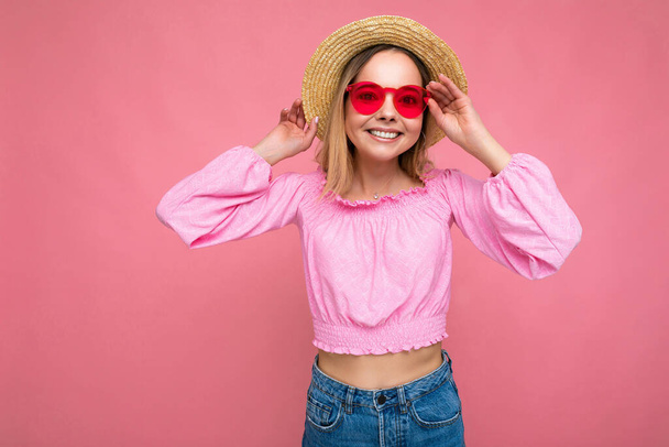Zdjęcie pięknej pozytywnej młodej blondynki noszącej letnie, luźne ubrania i stylowe okulary przeciwsłoneczne odizolowane od kolorowego tła patrzącego w kamerę - Zdjęcie, obraz