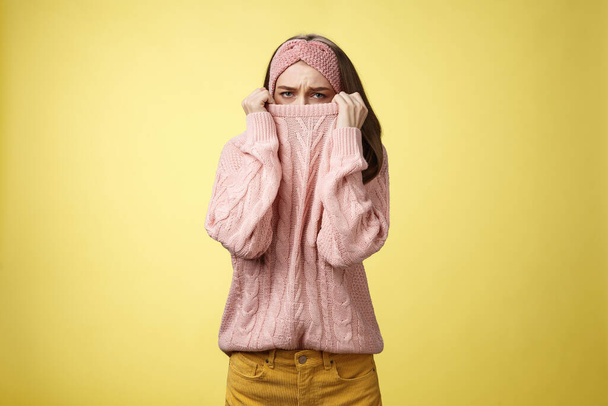 Sad silly, gloomy cute young girl hiding face in sweater collar pulling clothes on nose frowning displeased, unhappy, looking indignant and reluctant standing disappointed over yellow background - Foto, afbeelding