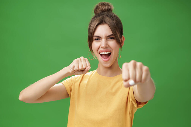 Mulher rebelde bonito em camiseta amarela com dentes lacuna puxando punho em direção à câmera como se mostrando habilidades de luta gritando ousado e animado em pé sobre fundo verde sorrindo agindo como boxeador - Foto, Imagem