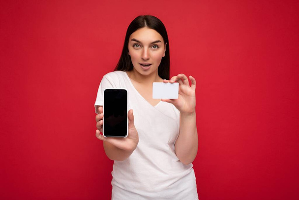 Foto de atractiva joven morena feliz con camiseta blanca casual aislada sobre fondo rojo con espacio vacío sosteniendo en el teléfono móvil de la mano y mostrando el teléfono inteligente con pantalla vacía para - Foto, Imagen