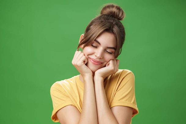 Tender sensual timid european girl in yellow t-shirt leaning head on shoulder touching cheeks with hands closing eyes and smiling with soft grin feeling nostalgic and romantic over green background - 写真・画像
