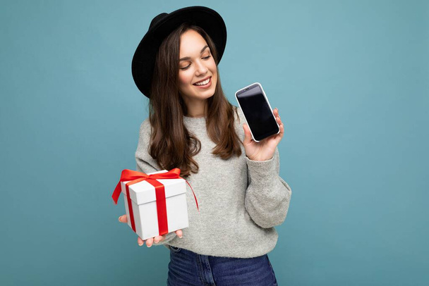 Φωτογραφία της όμορφης χαρούμενη νεαρή μελαχρινή γυναίκα απομονωμένη σε μπλε φόντο τοίχο φορώντας μαύρο καπέλο και γκρι πουλόβερ κρατώντας κουτί δώρου που δείχνει οθόνη του κινητού τηλεφώνου οθόνη για mockup και κοιτάζοντας - Φωτογραφία, εικόνα