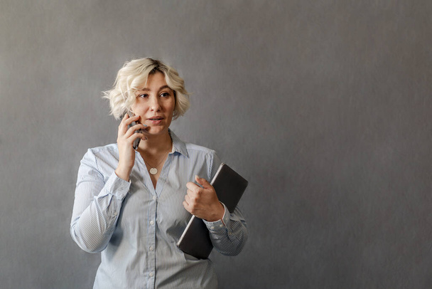 attractive young blonde talking on a mobile phone on a gray background, plus-size model, the concept of remote work online - Foto, imagen