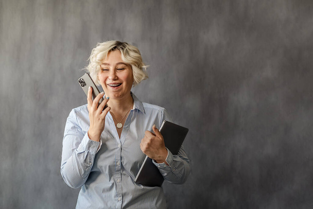 attractive young blonde talking on a mobile phone on a gray background, plus-size model, the concept of remote work online - Photo, Image