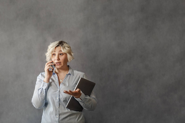 привлекательная молодая блондинка, разговаривающая на мобильном телефоне на сером фоне, плюс-размер модели, концепция удаленной работы в сети - Фото, изображение