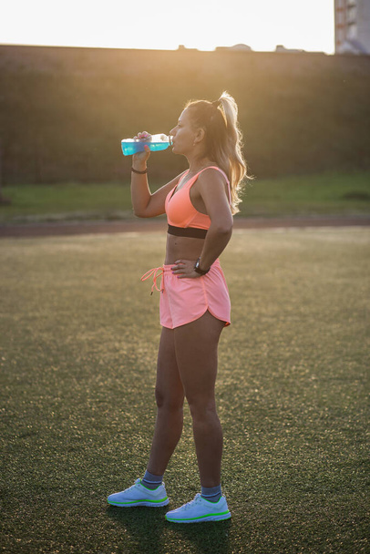 A woman is drinking water after training. A young and smiling girl is drinking a drink isotonic after physical fatigue. Female holds a bottle with blue liquid in her hand. Healthy lifestyle concept - Fotó, kép