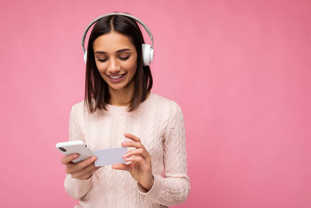 Photo shot of beautiful joyful smiling young female person wearing stylish casual outfit isolated over colorful background wall wearing white bluetooth wireless earphones and listening to music and - Zdjęcie, obraz