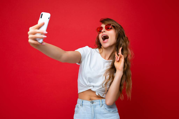 fotó csodálatos gyönyörű fiatal szőke nő gazdaság mobiltelefon fotózás szelfi fotó segítségével okostelefon kamera visel napszemüveg mindennapi elegáns ruhát elszigetelt át színes fal háttér látszó - Fotó, kép
