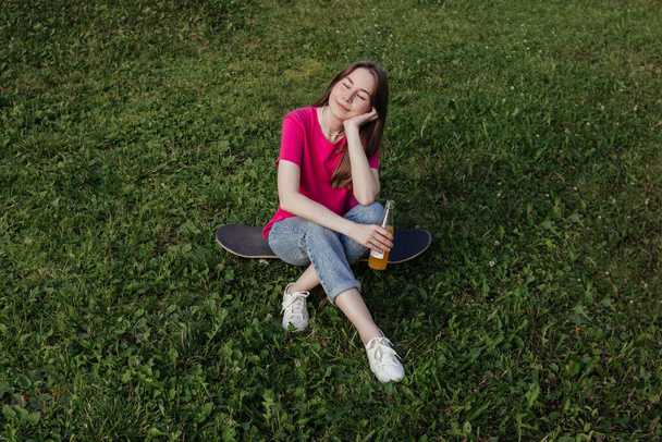 Young woman in pink t-shirt with closed eyes sitting on skateboard on green grass and holding bottle of lemonade. Summer activity, healthy lifestyle, sport, fitness. Female skater. Having fun outdoors - Photo, Image