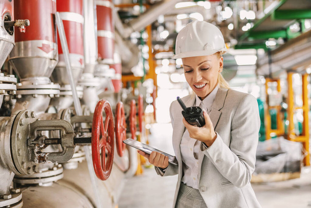 Middle-aged experienced hardworking female supervisor with helmet in suit holding digital tablet in hands and talking on walkies talkie with an employee while standing in the heating plant - Photo, Image