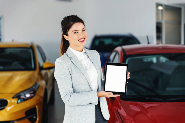 Przyjazna, uśmiechnięta kobieta dealerka samochodów stojąca w salonie samochodowym i pokazująca ekran tabletu. - Zdjęcie, obraz