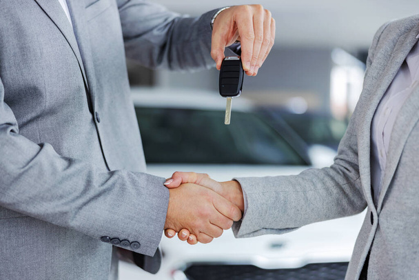 Closeup of car seller shaking hands with customer and handing her car keys while standing in car salon. - Photo, Image