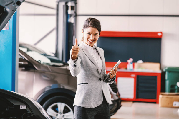 Female car seller standing in garage of car salon and showing thumbs up. Car is all set and repaired. - Фото, зображення