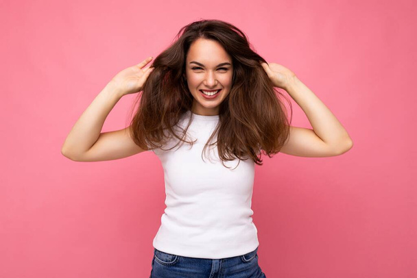 Portrait photo de jeune belle femme brune hipster souriante en t-shirt blanc avec maquette. Sexy insouciante femme posant isolé près du mur rose avec espace vide en studio. Modèle positif - Photo, image