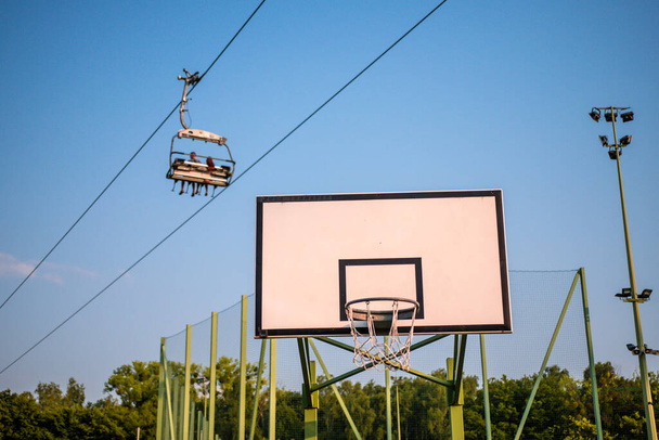 Basketbalové prkno proti vrcholkům stromů, modrá obloha a čtyřsedačková lanovka se třemi lidmi. - Fotografie, Obrázek
