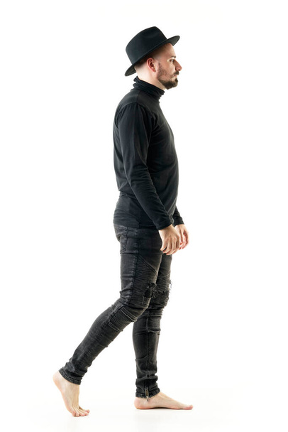 Zijaanzicht van artiest man performer in volledige zwarte kleren lopen blootsvoets. Full length portret geïsoleerd op witte achtergrond - Foto, afbeelding