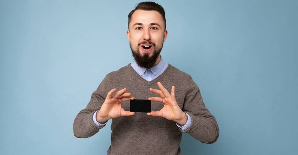 Panoramic photo of Handsome smiling brunette bearded guy wearing grey sweater and blue shirt isolated on background wall holding credit card looking at camera - Foto, imagen