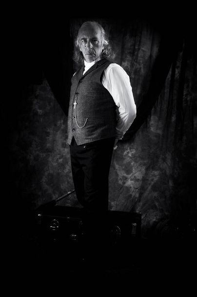 Czarno-biały, stojący portret dawnego księdza stojącego w ciemnym pokoju z rękami za plecami, gapiącego się na widza. - Zdjęcie, obraz