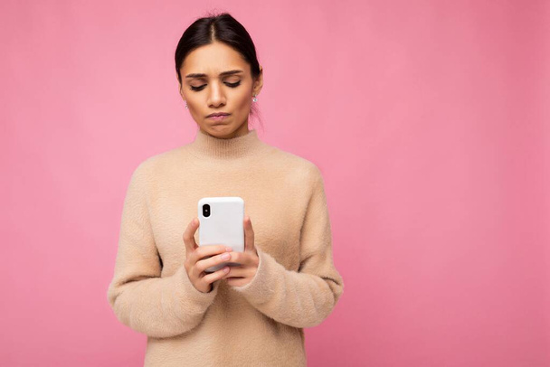 atraente bonito chateado jovem morena mulher vestindo bege quente suéter de pé isolado sobre rosa fundo surf na internet via telefone olhando para a tela móvel - Foto, Imagem