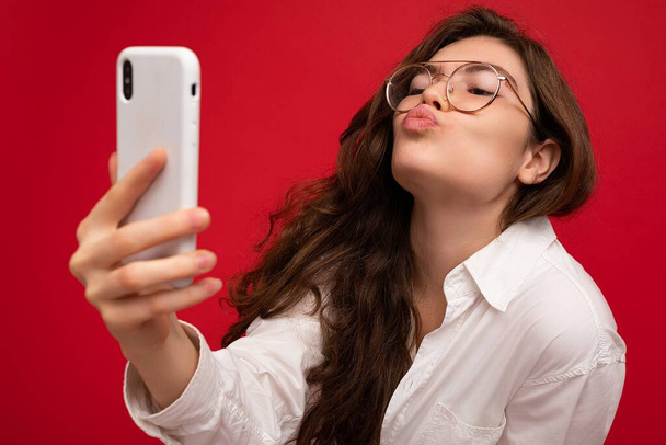 Szoros fotó szexi gyönyörű pozitív fiatal barna nő visel fehér ing és optikai szemüveg elszigetelt felett piros háttér gazdaság a kezében, és a mobiltelefon segítségével selfiie nézi - Fotó, kép