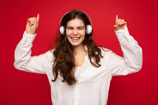 Atraktivní šťastný úsměv mladá brunetka žena osoba nosí bílou košili a optické brýle izolované přes červené pozadí na sobě bílé bezdrátové bluetooth sluchátka poslech hudby a tance - Fotografie, Obrázek