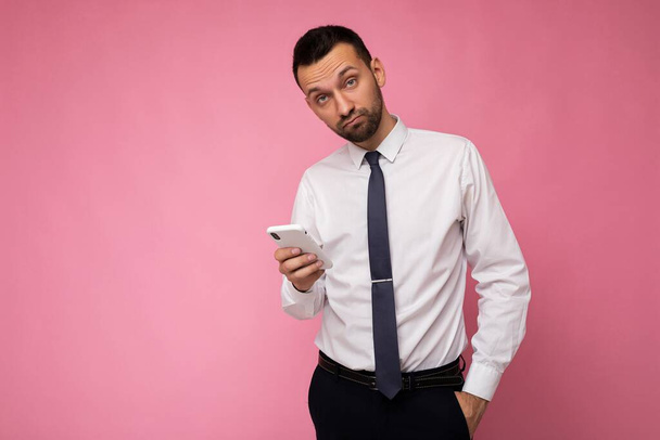 Fénykép jóképű jóképű férfi visel alkalmi fehér ing és nyakkendő elszigetelt rózsaszín háttér üres hely tartja a kezében, és a mobiltelefon üzenetküldő sms nézi a kamerát - Fotó, kép