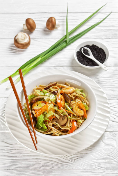 Prawn Yaki Udon, stir fried udon noodles with shrimps, cabbage, chestnut mushrooms, spring onion, carrot, mirin and soy sauce in white bowl with chopsticks,  japan cuisine, vertical view - Photo, image