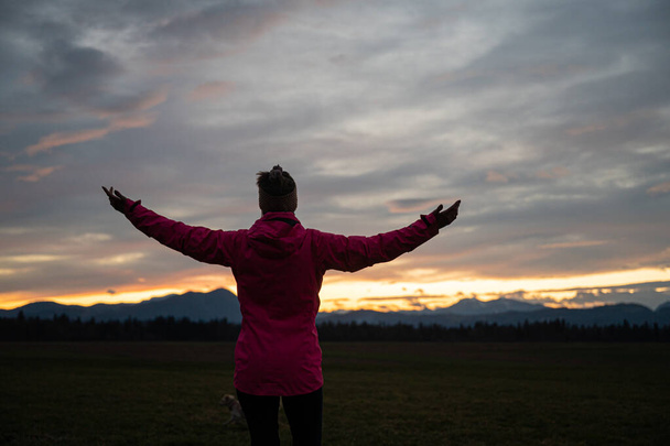Vista da dietro di una giovane donna in giacca rosa in piedi con le braccia aperte ampiamente sotto un cielo serale nuvoloso. - Foto, immagini