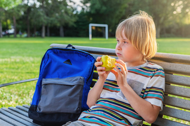 Pequena criança estudante do ensino fundamental sentada no banco perto do pátio da escola com mochila, tendo lanche após a escola ou durante o recreio. - Foto, Imagem