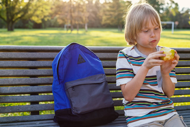 Kleine basisschoolstudent zittend op een bankje in de buurt van het schoolplein met rugzak, snack na schooltijd of tijdens de pauze. Terug naar school. - Foto, afbeelding