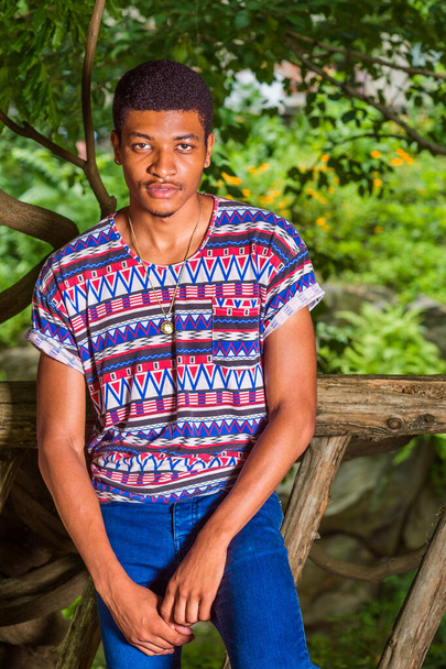 Portret van de jonge zwarte man. Het dragen van een kleurrijk patroon shirt, blauwe jeans, ketting, een jonge knappe man staat tegen een houten hek, charmant kijken naar je - Foto, afbeelding