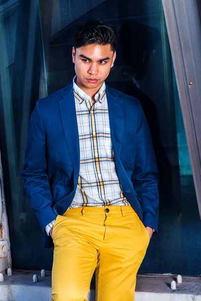 Der Mensch denkt im Freien. Im blauen Blazer, Unterwäsche im Muster, gelbe Hose, Hände in Taschen, steht ein junger Mann vor einer Glaswand auf der Baustelle, senkt den Kopf, denkt nach. - Foto, Bild