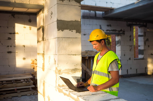 Construction engineer on the construction site of a house made of porous concrete blocks works at a computer in a reflective safety vest and hardhat. Design, construction, drawings project, check - Zdjęcie, obraz
